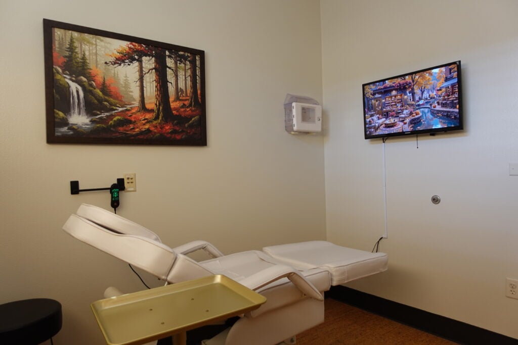 Private treatment room equipped with a white reclining medical chair, sanitation station, and wall-mounted TV.